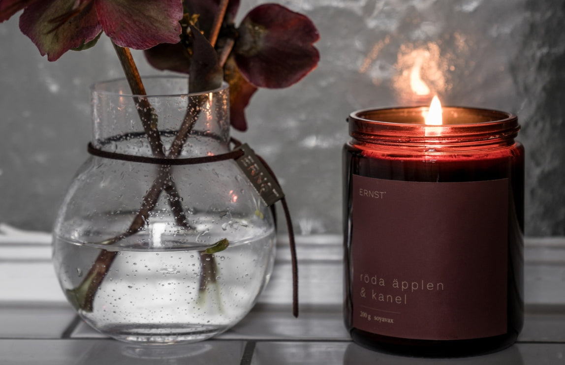 Doftljus röda äpplen från Ernst i röd glasburk, bredvid står en vas med röda blommor- Bild till kategorin Doftljus och Doftpinnar