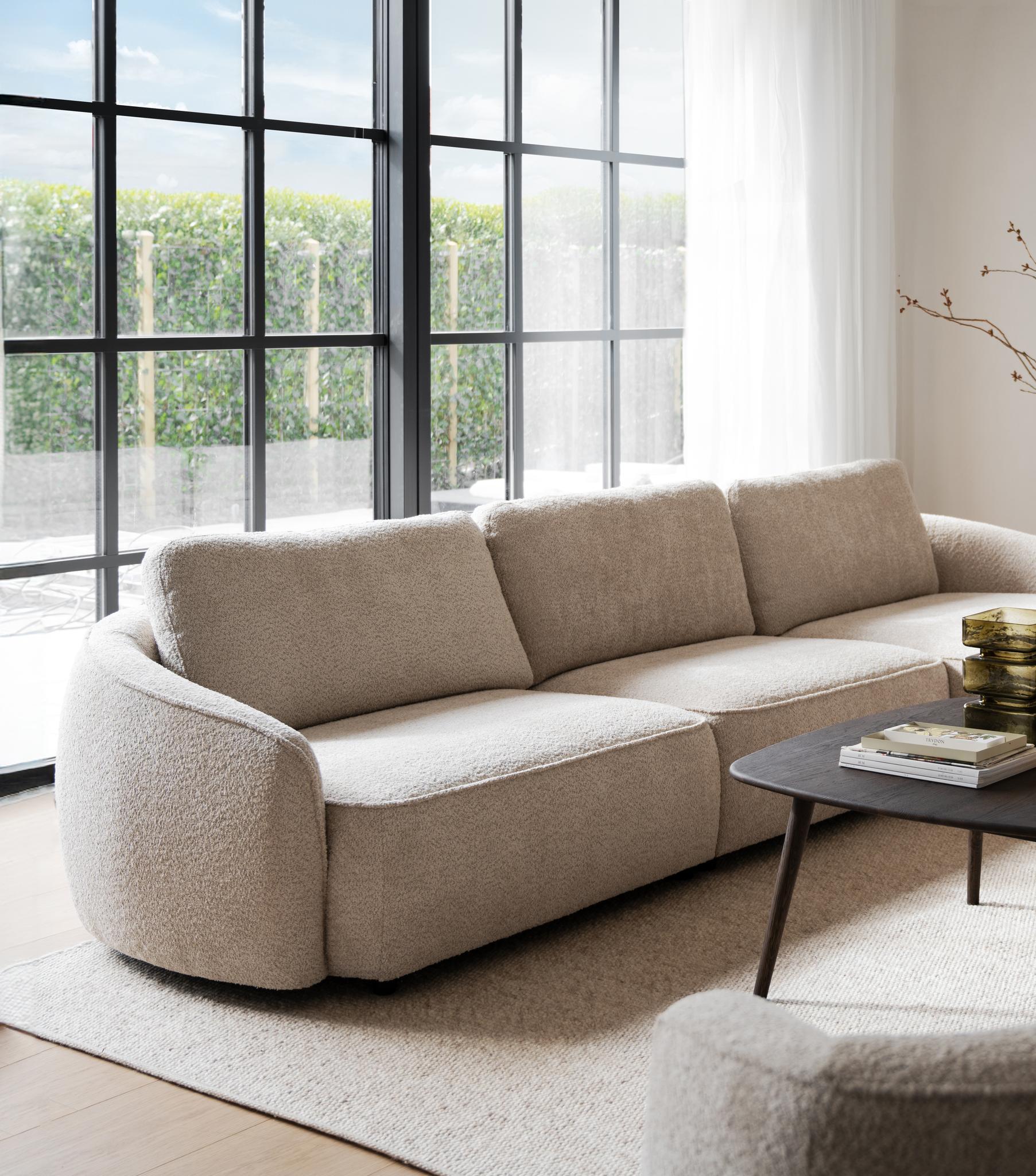 Beige bouclésofa med rundade former i ljust vardagsrum med stora fönster och minimalistisk inredning