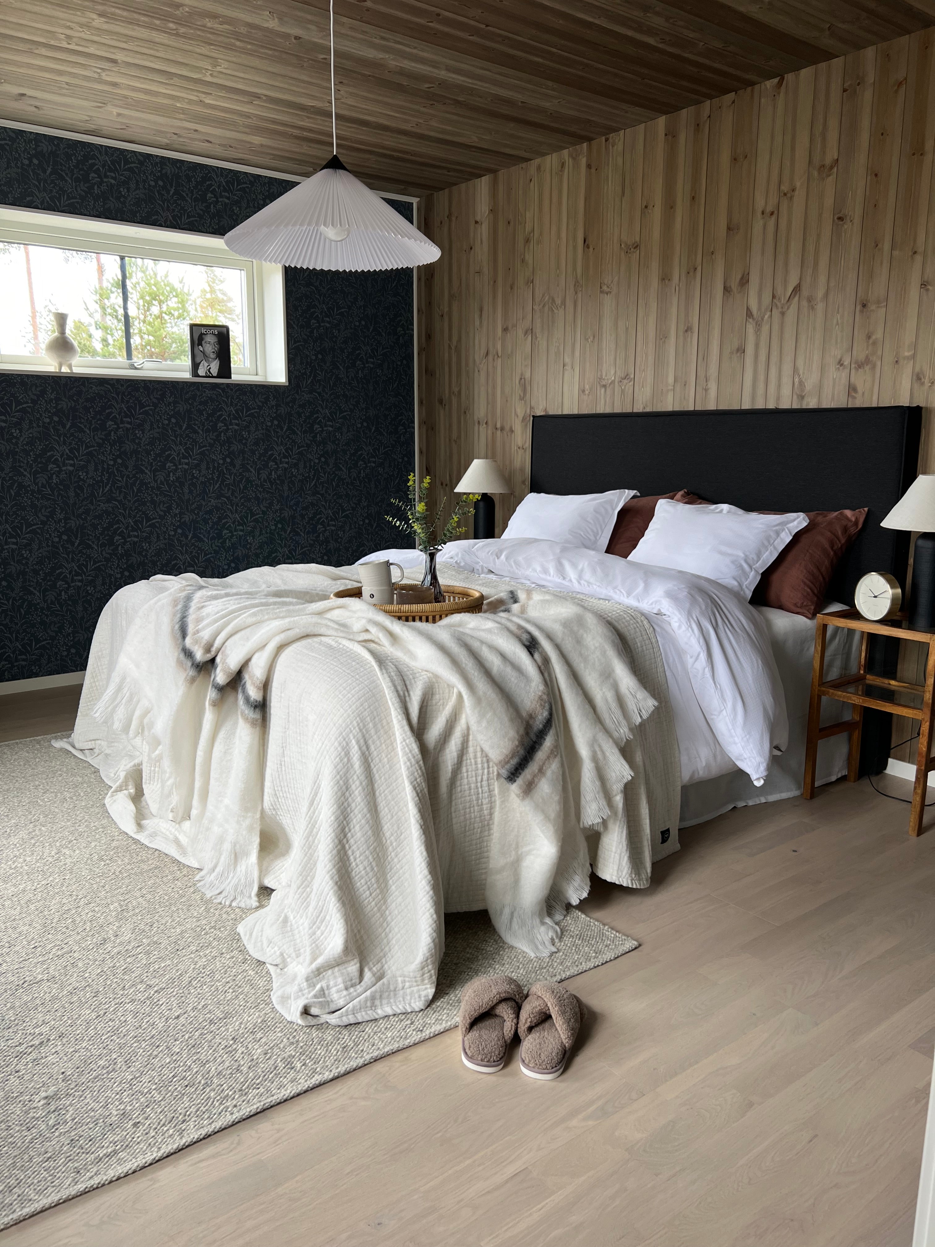 Mysigt sovrum med ljust trägolv, sängkläder i vitt och beige, svart sänggavel och dekorativ taklampa