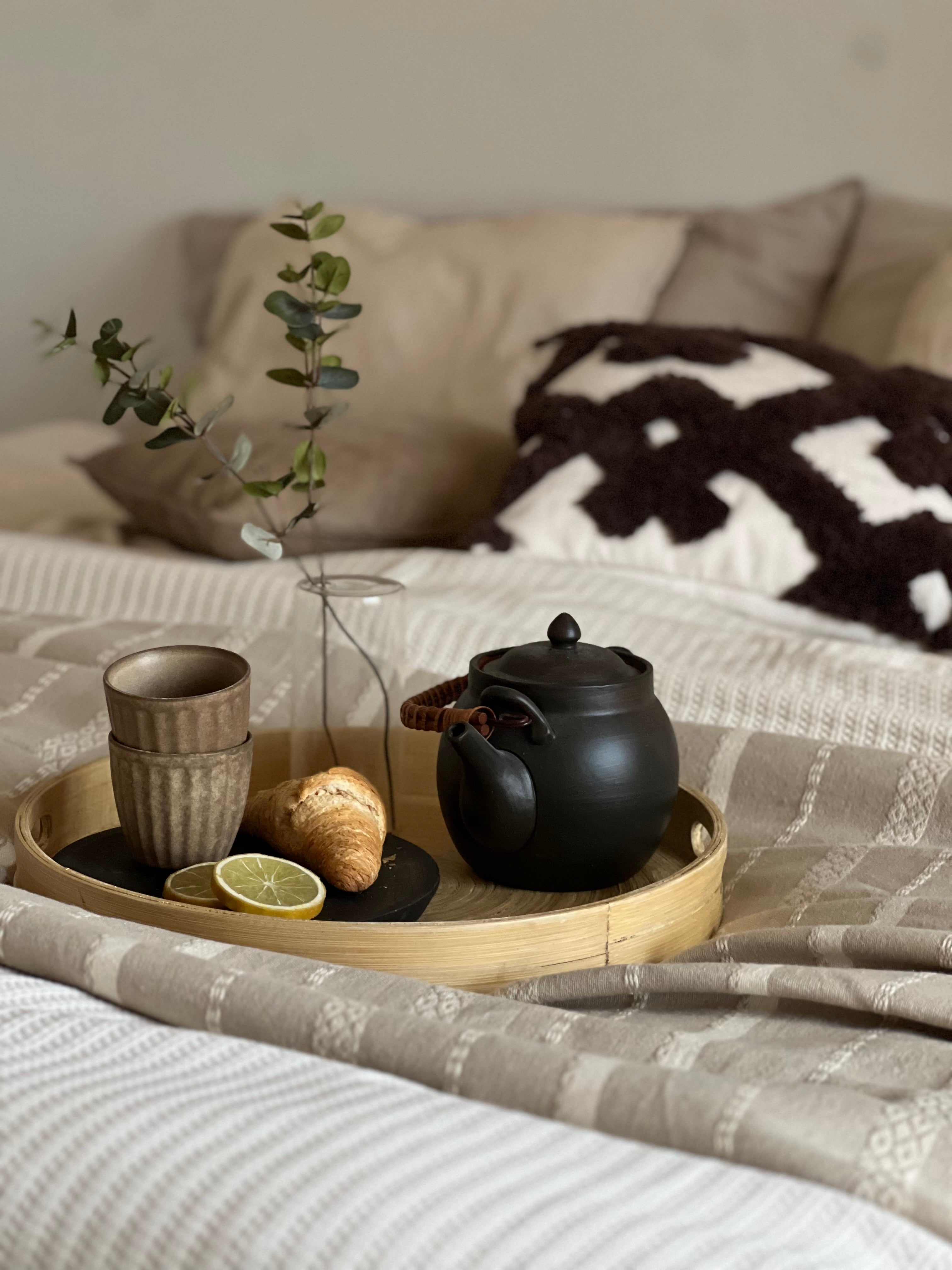 Frukostbricka i bambu med tekanna, croissant och citron på sängen med dekorativa kuddar