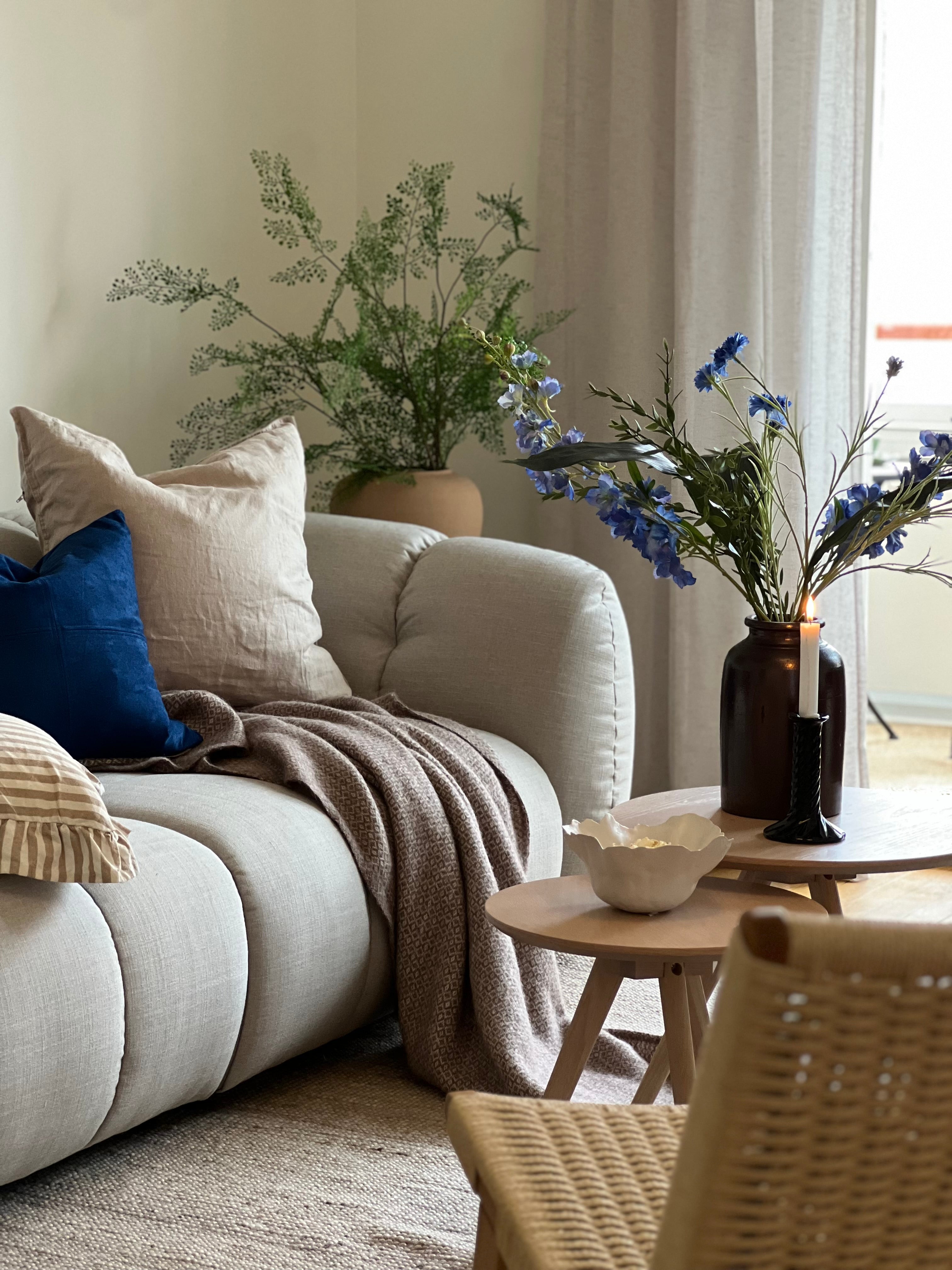 Ljust vardagsrum med beige sammetssoffa, blå och vita kuddar, brun pläd och blommor i vas på soffbord