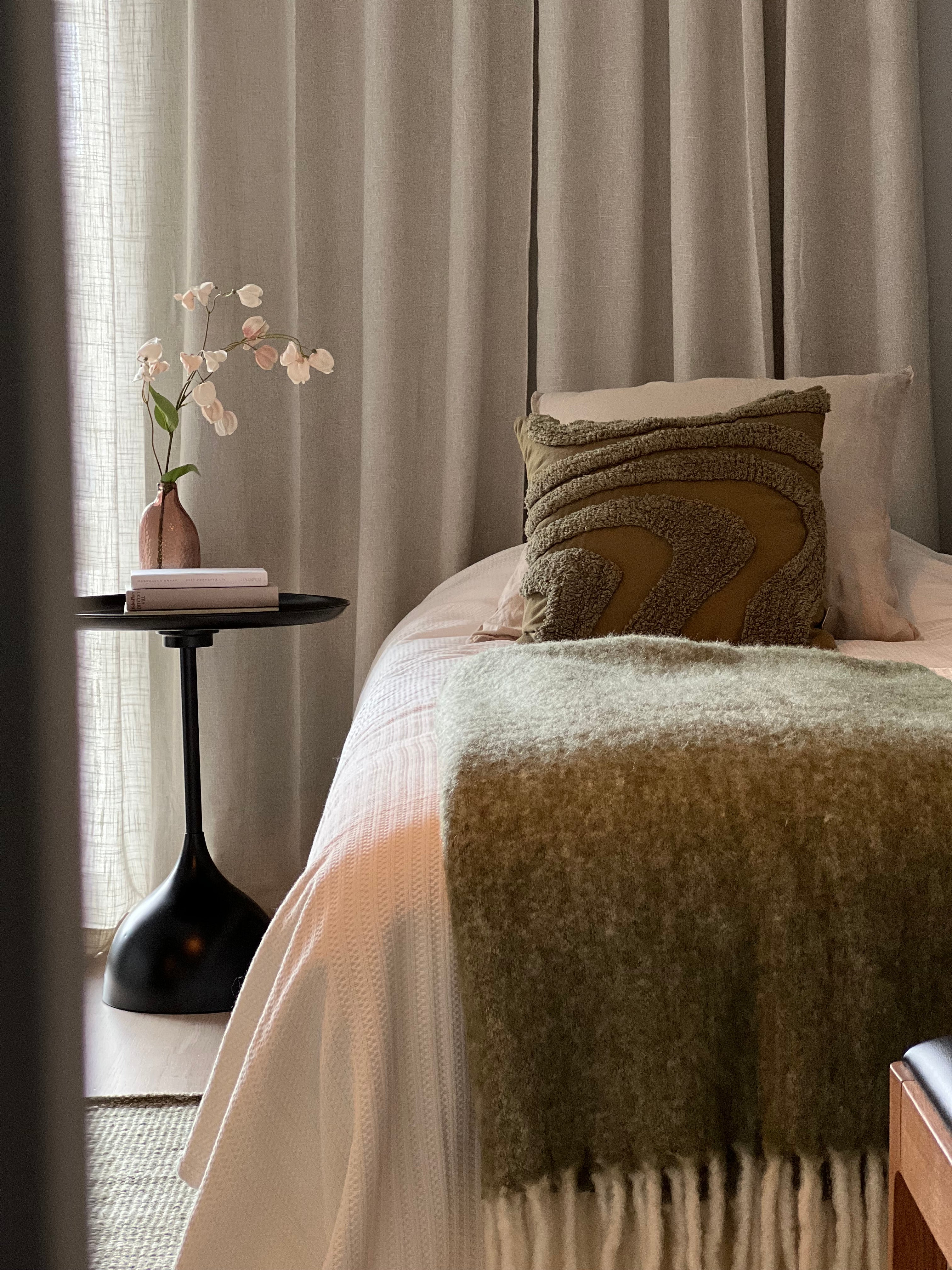 Sovrum med beige sängkläder, olivgrön pläd med fransar och dekorativ kudde bredvid svart sidobord