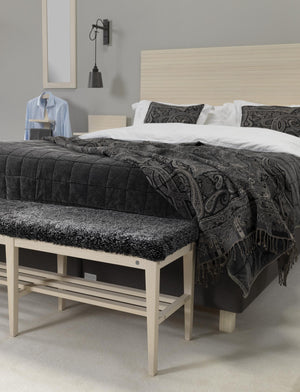 Sovrum med ljus träsäng, vit sängkläder, mörk överkast med paisleymönster och grå vägglampa