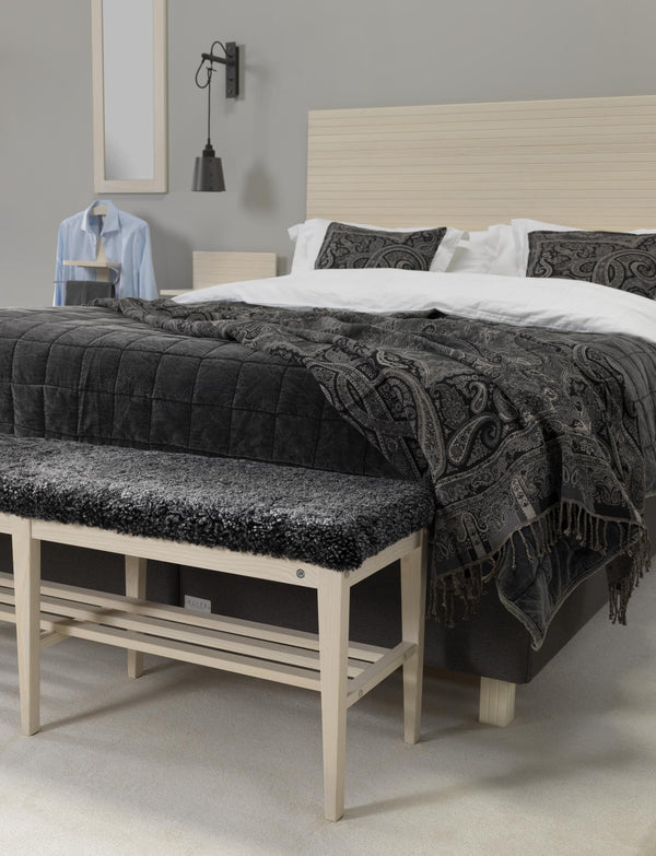 Sovrum med ljus träsäng, vit sängkläder, mörk överkast med paisleymönster och grå vägglampa