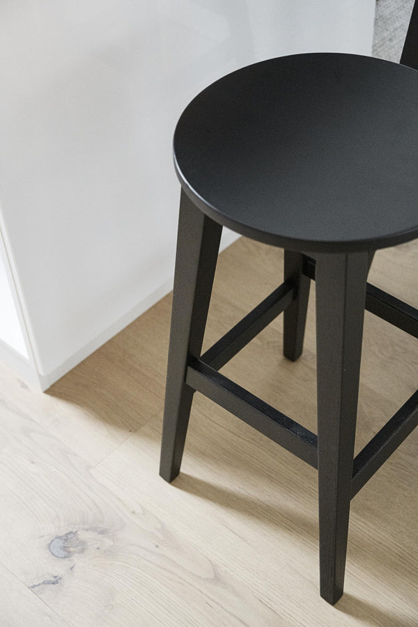 Svart rund pallfåtölj med träben står på ljust trägolv intill vit vägg