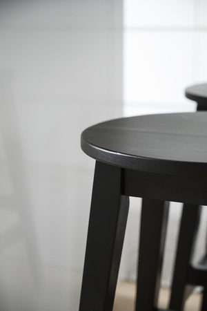 Svart träpall med rundad sits och fyra ben i modern minimalistisk design mot ljus vägg