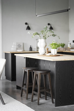 Modernt kök med svarta skåp, ljus träbänkskiva, barpall och gröna växter i vit vas