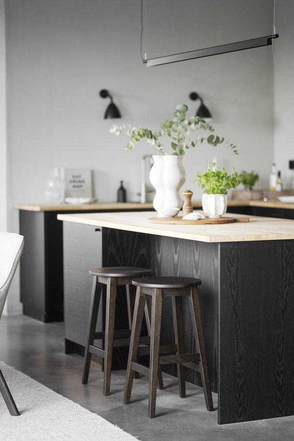 Modernt kök med svarta skåp, ljus träbänkskiva, barpall och gröna växter i vit vas