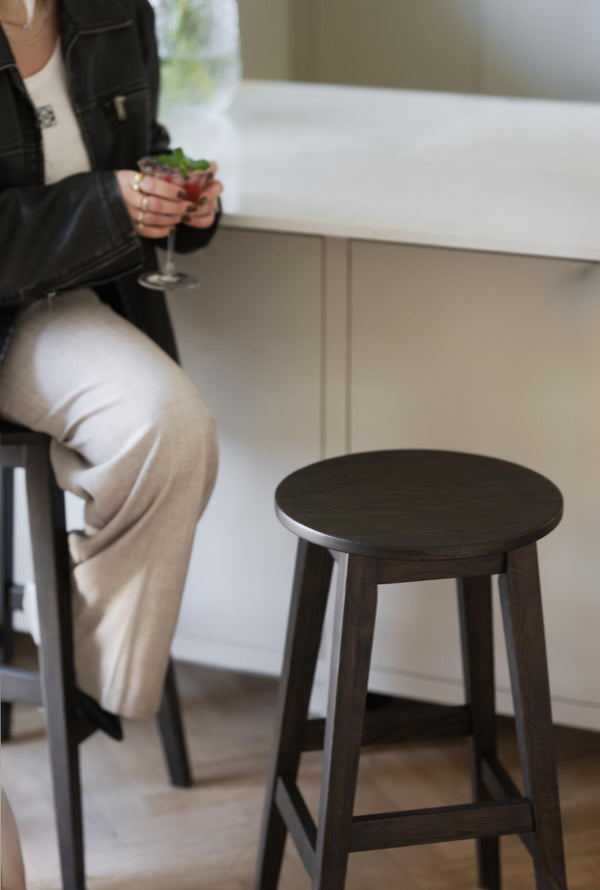 Kvinna sitter på mörk träpall vid diskbänk med drink i handen, vit köksinredning i bakgrunden