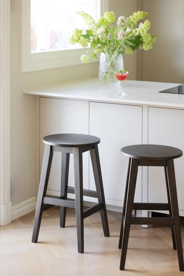 Två mörkbruna barstolar i trä framför vit köksbänk med blommor och drinkglas