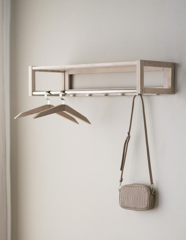Väggmonterad hallhylla i ljust trä med krokar, två galgar och en beige väska med axelrem