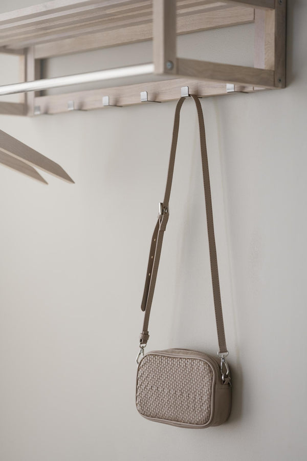 Beige crossbody-väska i flätat material hängande på krok under hyllsystem i ljust trä
