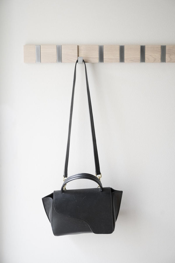 Svart handväska hängande på krok i trälist med metalldetaljer mot vit vägg