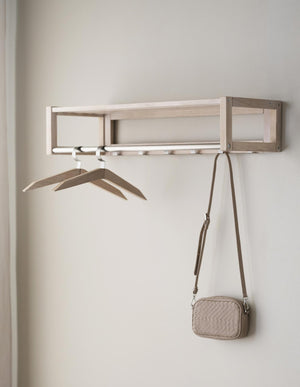 Väggmonterad hattshylla i ljust trä med krokar, två galgar och en beige väska hängande