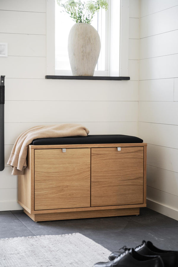 Hallbänk i ek med svart dyna och förvaringslådor, beige pläd, vit vägg med hylla och vas