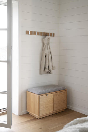 Hallbänk i ek med förvaringsskåp och grå dyna, väggmonterad krokplanka med kardigan i minimalistisk hall
