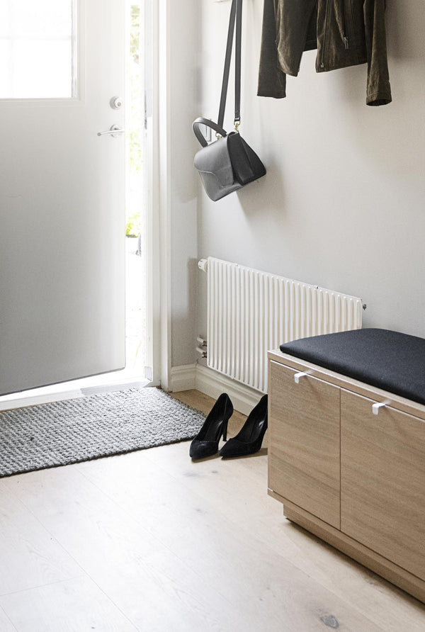 Minimalistisk hall med bänk i ljust trä, grå matta, svarta skor och väska hängande på väggen