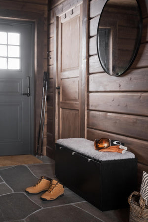 Välkomnande hall med träpanel, rund spegel, svart bänk med sits och bruna vinterkängor på stengolv