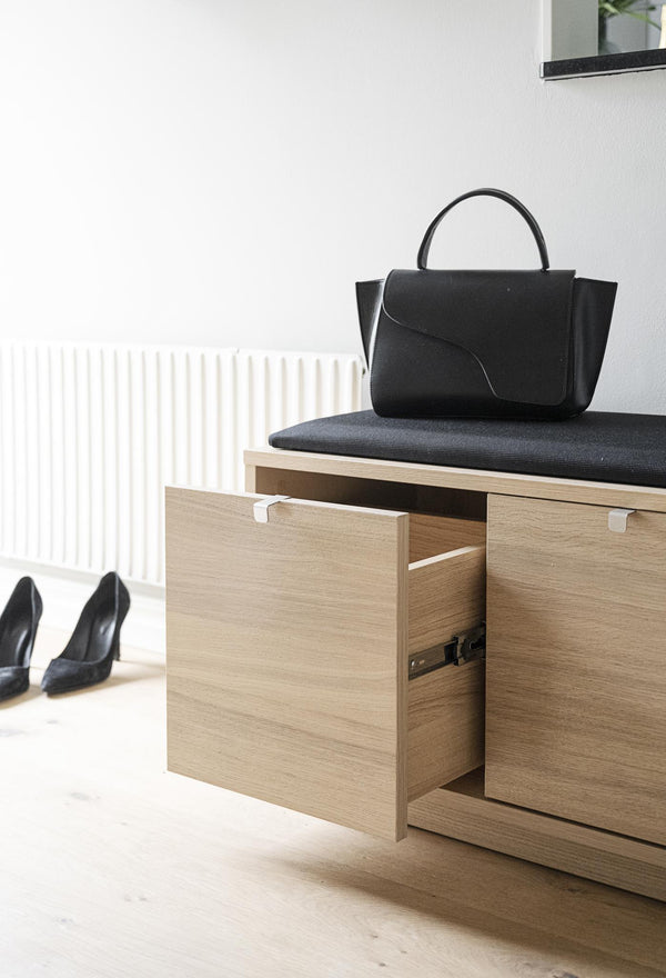 Hallbänk i ek med öppen låda, svart stoppning och svart väska, med skor framför vit vägg