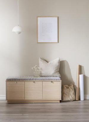 Ljust hallmöbel i trä med tre dörrar, grå dyna och kudde, vit lampa och inramad tavla på väggen