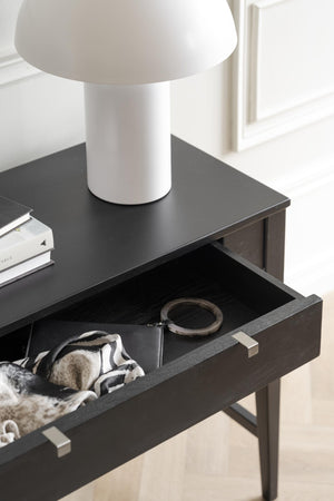 Svart sängbord med utdragen låda innehållande smycken och accessoarer, vit bordslampa ovanpå