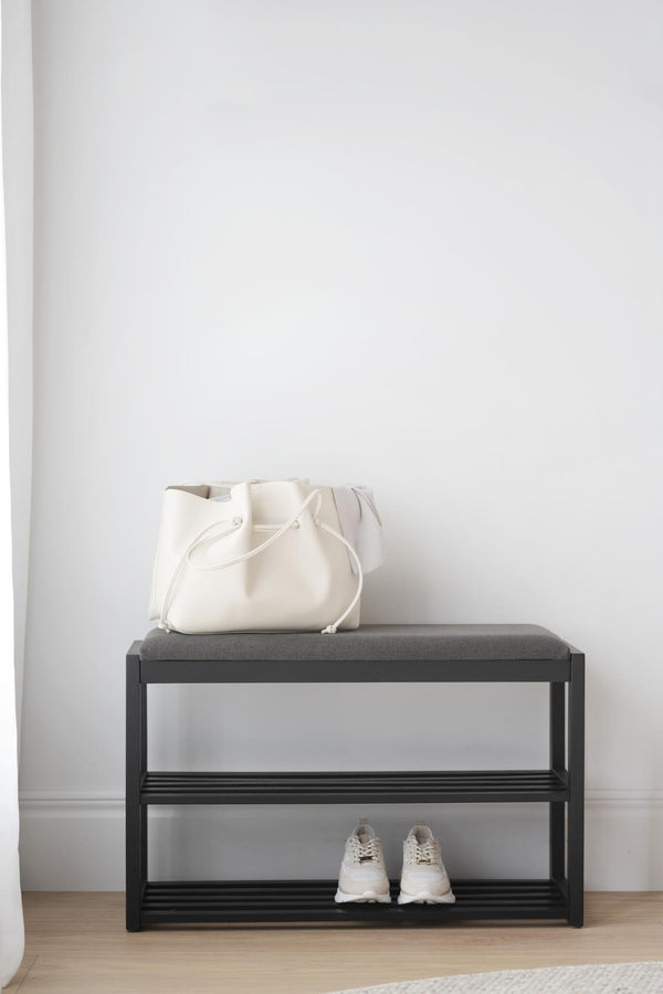 Skoförvaring med bänk i svart metall med grå dyna, vit väska och vita sneakers i minimalistisk hall