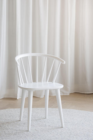 Vit pinnstol i trä med spjälor i ryggen, stående på ljus matta framför vita gardiner