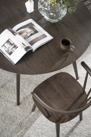Runt matbord i mörk ek med matchande stol, dekorerat med tidning, kaffekopp och vas med gröna kvistar