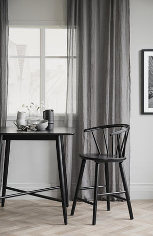 Svart matbord med stol och grå gardiner vid fönster i minimalistisk matsal med trägolv