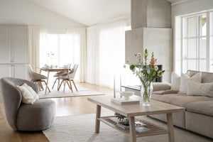 Ljust vardagsrum med beige soffa, soffbord i trä, vita gardiner och matplats i bakgrunden