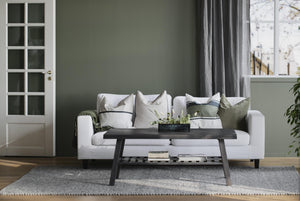 Vit soffa med kuddar, mörkt soffbord och grå matta i ljust vardagsrum med gröna väggar