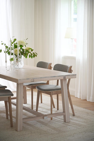 Matbord i ljust trä med gråa stoppade stolar i ljust matrum med vita gardiner