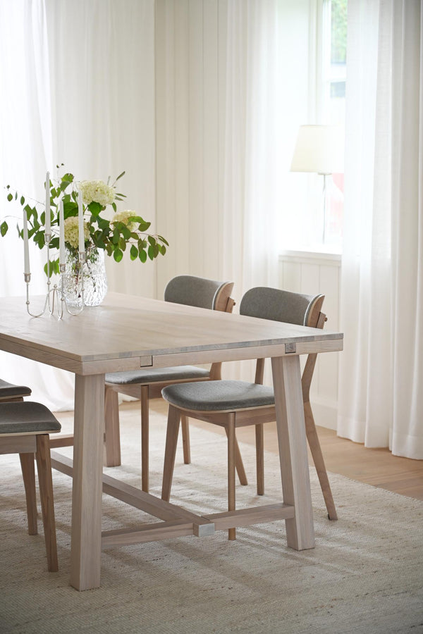 Matbord i ljust trä med gråa stoppade stolar i ljust matrum med vita gardiner