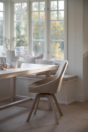 Beige matstol med armlän vid ljust matbord framför fönster med sittplats och vita kuddar