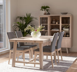 Ljus matgrupp i trä med sex grå stoppade stolar, vitrinskåp och växter i skandinavisk matsal