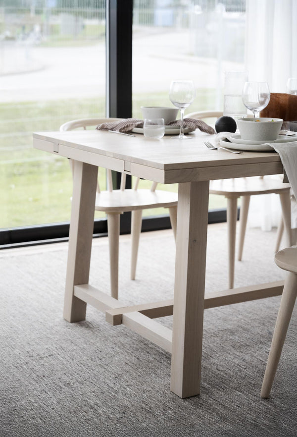 Vitt matbord i trä med fyra ben och hyllan undertill, dukat med tallrikar och glas vid fönster