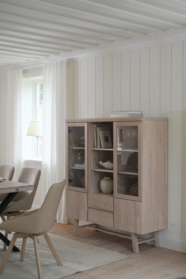 Vitrinskåp i ljust trä med glasdörrar och lådor i skandinavisk matsal med vita panelväggar