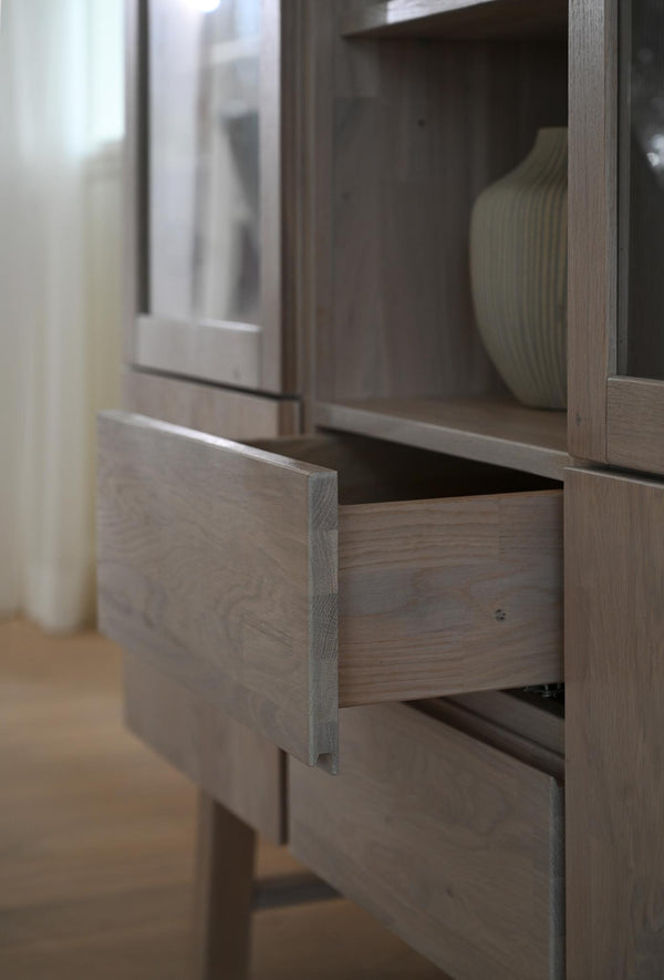 Öppen låda i vitrinskåp av ljust trä med glasdörrar och synlig grön keramikvas