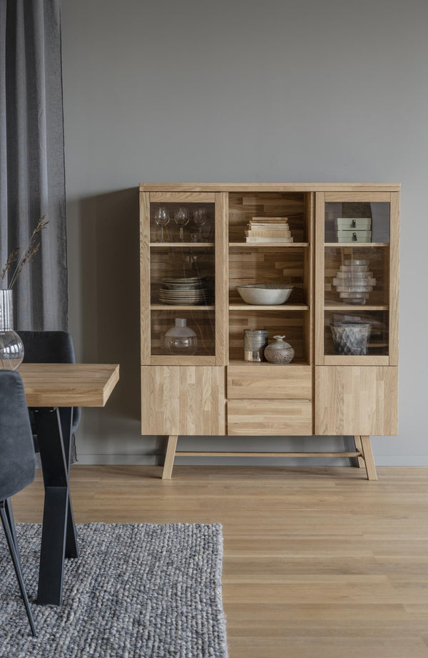 Vitrinskåp i ljust trä med glasdörrar, öppna hyllor och lådor, fyllt med porslin och inredningsdetaljer