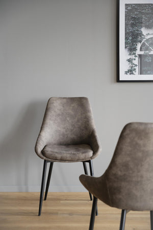 Gråbrun matstol i sammet med svarta ben på trägolv mot vit vägg med tavla