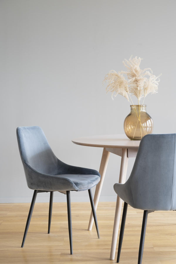 Grå sammetsstolar med svarta ben vid runt matbord i ljust trä med glasvas och torkade blommor