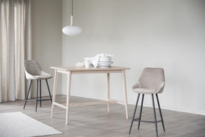 Matbord i ljust trä med hylla, två grå barstolar och vit taklampa i minimalistisk matsal