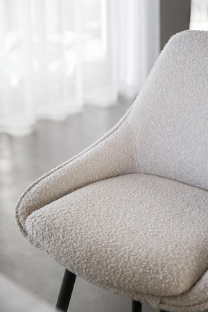 Närbild på beige fåtölj med bouclétyg och svarta ben vid fönster med vita gardiner