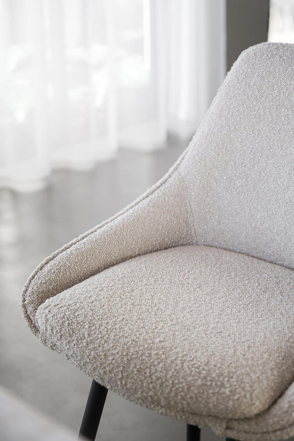 Närbild på beige fåtölj med bouclétyg och svarta ben vid fönster med vita gardiner