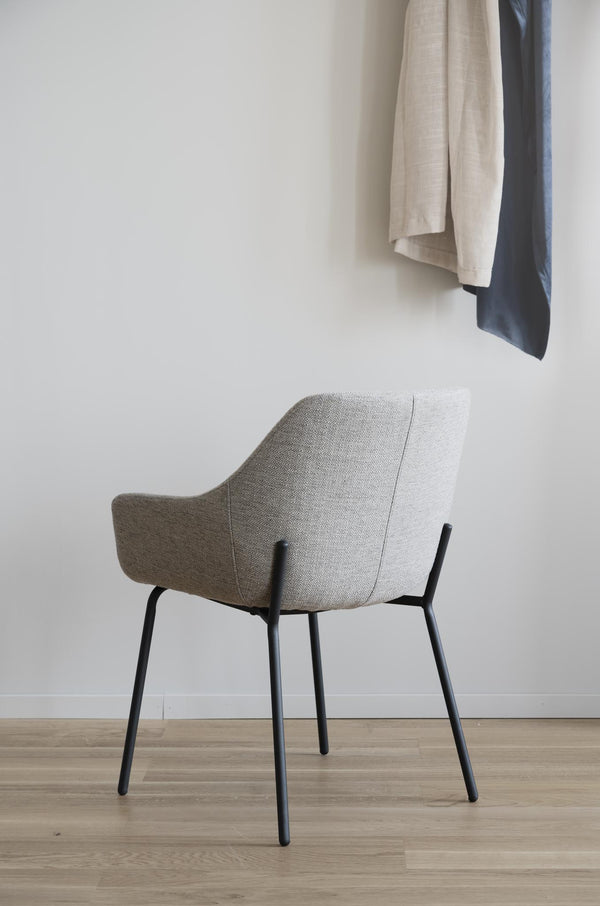 Grå stol med armstöd och svarta ben i modern skandinavisk design mot vit vägg med hängande kläder