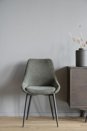 Grön stoppad matstol med svarta metallben vid mörkbrun skänk med vas och torkade blommor