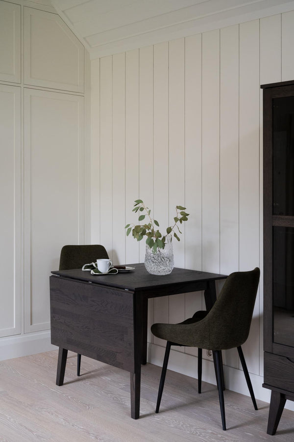 Svart matbord med två gröna stolar mot vit panelvägg i skandinavisk stil