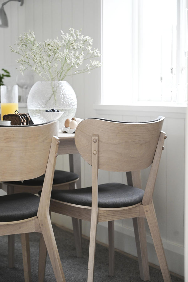 Ljusa trämatbordsstolar med svart sits vid runt matbord med vitfrostat glasvas och blommor