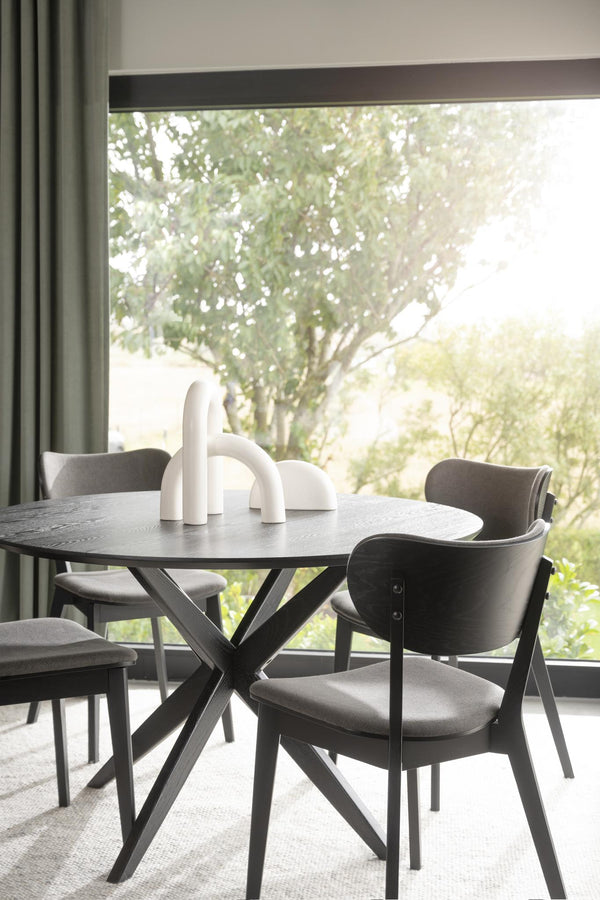 Runt matbord i mörkt trä med korsben och grå stolar vid fönster med utsikt mot grönska