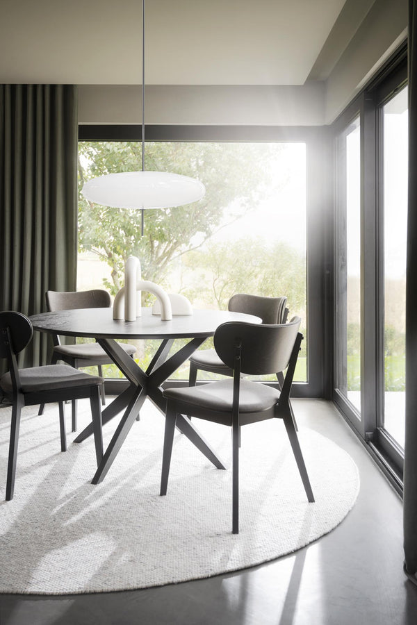 Rund matbord med korsben i svart och grå stolar i modern matsal med stora fönster och utsikt mot grönska